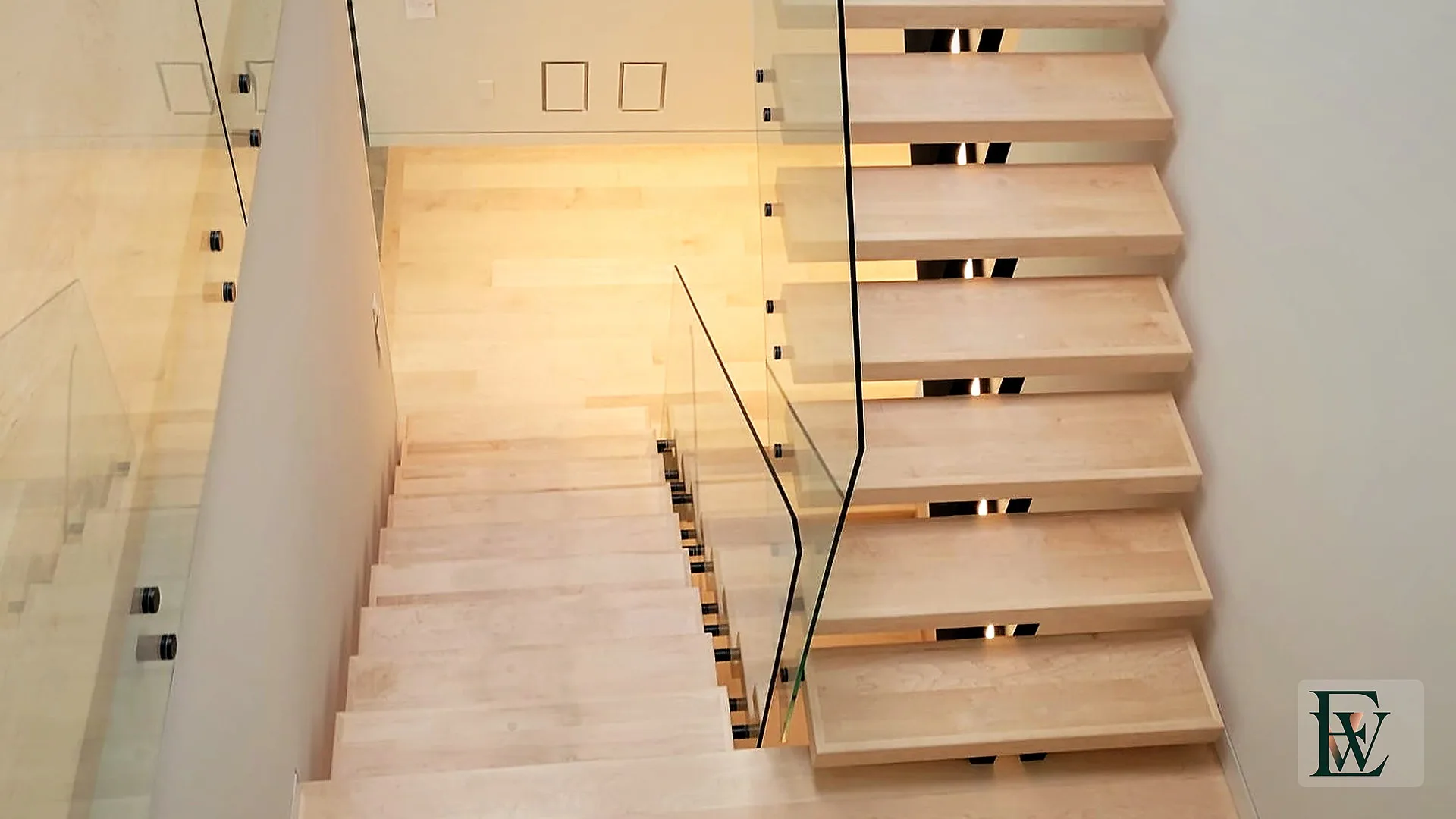 Select hard maple stair treads and landing seen from the upper void, framed by frameless glass guardrails.